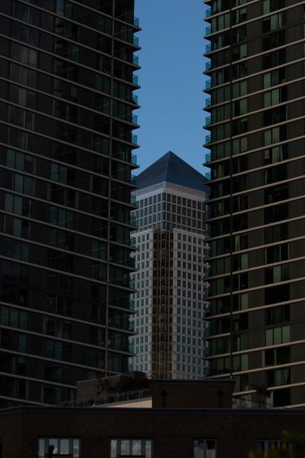 The image depicts a high-angle view of a modern urban garden balcony in Canary Wharf, framed by two tall residential or office buildings with dark glass facades featuring horizontal balcony railings. 
