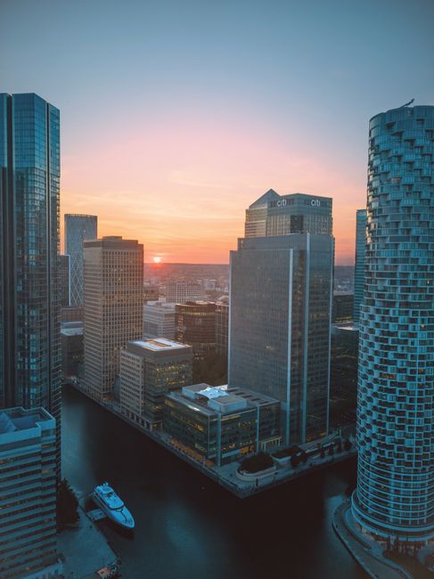 The image depicts a high-angle view of a modern urban garden balcony in Canary Wharf, framed by two tall residential or office buildings with dark glass facades featuring horizontal balcony railings. 