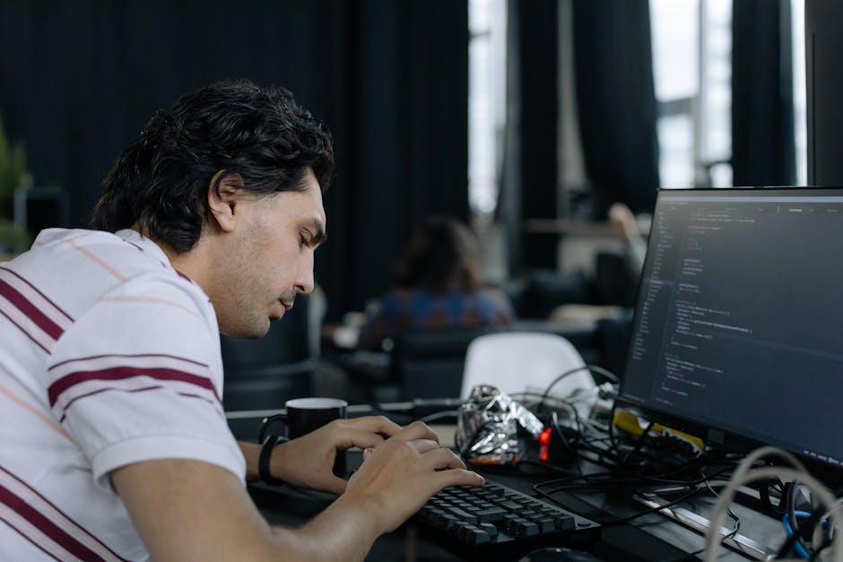 A young man with dark, curly hair and light skin is working at a desk in a modern office environment, focused on a computer screen displaying lines of code. He is wearing a white shirt with horizontal